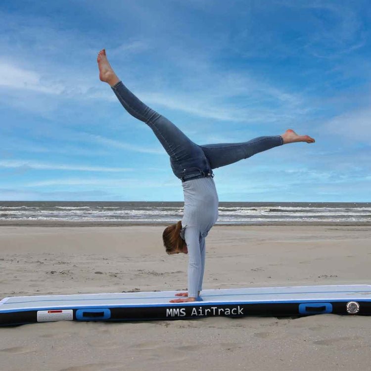 Een vrouw maakt een handstand op de MMS AirTrack Turnmat op het strand, met de zee en een blauwe lucht op de achtergrond.